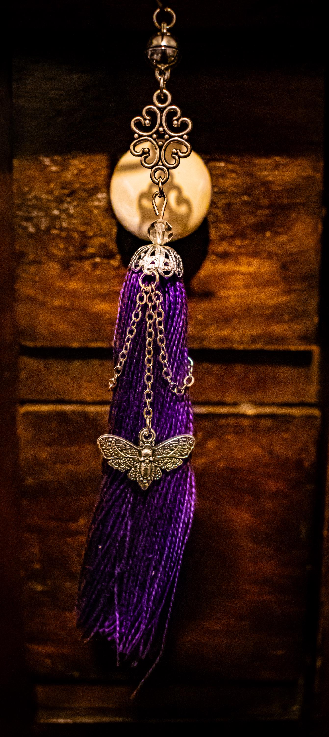 Bookmark tassel with purple threads, silver chains, a moth and skull charm hanging from a silver chain, and a silver charm made of filigree connected to the top of the tassel.