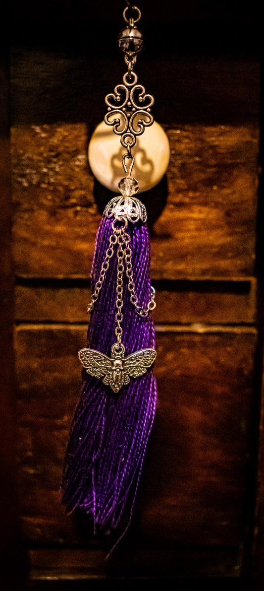 Bookmark tassel with purple threads, silver chains, a moth and skull charm hanging from a silver chain, and a silver charm made of filigree connected to the top of the tassel.